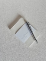 A textured cream-colored surface serves as the backdrop for a notebook positioned slightly open, revealing a blank page. Resting on top of the notebook is a black and silver pen. The notebook cover displays text that reads 'Slowords One Day, A Day, One by Johnnie Santon'.