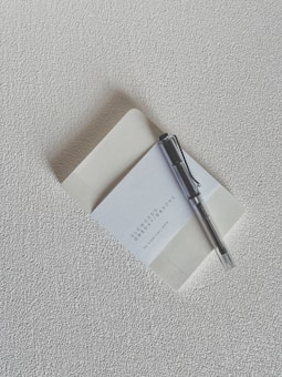 A textured cream-colored surface serves as the backdrop for a notebook positioned slightly open, revealing a blank page. Resting on top of the notebook is a black and silver pen. The notebook cover displays text that reads 'Slowords One Day, A Day, One by Johnnie Santon'.