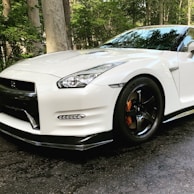 A sleek white sports car with a glossy finish, featuring sharp lines and aerodynamic design, parked on a textured asphalt surface. The car has a prominent front grille, detailed headlights, and black alloy wheels with silver brake calipers. The background includes a lush green setting with trees.