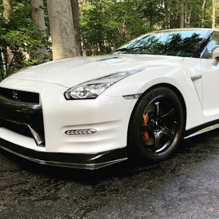 A sleek white sports car with a glossy finish, featuring sharp lines and aerodynamic design, parked on a textured asphalt surface. The car has a prominent front grille, detailed headlights, and black alloy wheels with silver brake calipers. The background includes a lush green setting with trees.