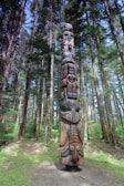 A tall, intricately carved totem pole stands in the midst of a dense, lush forest. The totem is adorned with various animal and human motifs, showcasing detailed craftsmanship. Surrounding the totem are towering trees with abundant green foliage, and sunlight filters through the branches, casting dappled light on the ground.