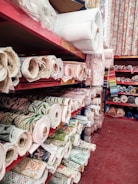 Multiple shelves are filled with rolled up fabric in various patterns and colors. The shelves are organized and display a variety of textures and designs suitable for upholstery or sewing projects. The floor is a deep red color, and some fabrics feature floral and geometric patterns.