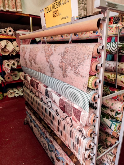 A display of various rolls of fabric on a rack, with patterns ranging from a world map to geometric and floral designs. A yellow sign above reads 'TELA RESINADA 6.50€ 1.40 ANCHO'. The setting appears to be a fabric store with shelves filled with different types of fabric.