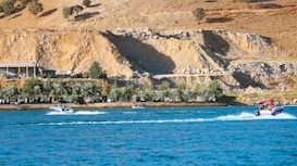 Two motorboats speed across a vibrant blue body of water, leaving white trails behind them. In the background, a rugged hillside with sparse vegetation and rocky terrain rises above a row of lush green trees lining the water's edge. A single-story building is visible on the left side of the image, partially obscured by trees.