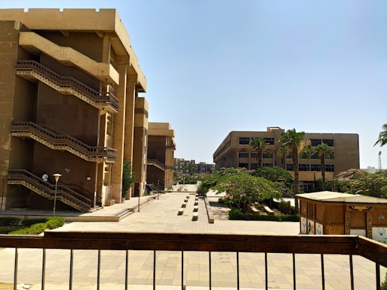 A campus area featuring tall, beige-colored buildings with staircases on the exterior. The open courtyard is lined with palm trees and other green foliage. A bright, clear sky is visible above, and the area is well-lit by sunlight. The ground is made of neatly laid tiles, and the setting is calm and orderly.