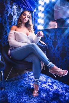 A woman is sitting in a stylish, plush room with decorative blue wallpaper and carpet. She is seated on a chair, wearing a white off-shoulder top, blue jeans, and white strappy heels. The setting includes a mirror with bright lights, creating a glamorous and high-fashion atmosphere.