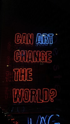 Bright neon lights display the phrase 'CAN ART CHANGE THE WORLD?' in a combination of red and blue colors, set against a dark background.