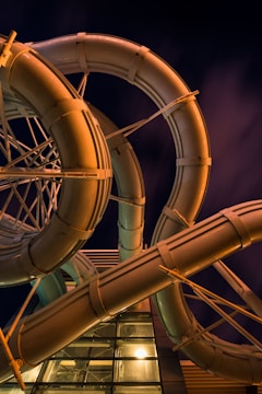 Curving water slides intertwined with each other at a nighttime setting, illuminated by artificial lights that give them a warm glow. The background is a deep, dark sky with subtle cloud formations.