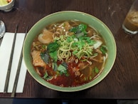 A bowl of Vietnamese noodle soup is garnished with fresh herbs and chili paste, served on a wooden table alongside chopsticks and a napkin. The soup contains slices of meat and is paired with a glass of iced drink.