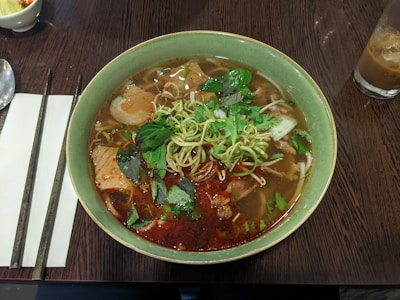 A bowl of Vietnamese noodle soup is garnished with fresh herbs and chili paste, served on a wooden table alongside chopsticks and a napkin. The soup contains slices of meat and is paired with a glass of iced drink.