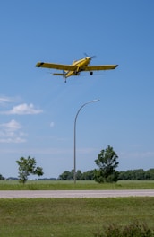 A yellow agricultural plane flies low over a grassy field, with trees and a single street lamp visible in the background under a clear blue sky.