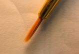 A close-up of a fine-tipped paintbrush with an orange bristle, set against a soft, beige background. The metal ferrule holding the bristles is gold, adding a touch of sophistication.