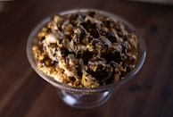 A glass bowl is filled with popcorn drizzled with rich chocolate and caramel. The popcorn appears glossy and decadent, sitting on a dark wooden surface which adds contrast to the presentation.