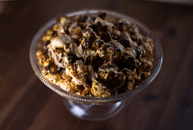 A glass bowl is filled with popcorn drizzled with rich chocolate and caramel. The popcorn appears glossy and decadent, sitting on a dark wooden surface which adds contrast to the presentation.