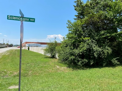 A street sign labeled 'Science.gov Way' stands in front of a grassy area beside a road. To the right, there is a large tree with dense green foliage. In the background, an industrial-looking building with a metal roof is visible beneath a clear blue sky.