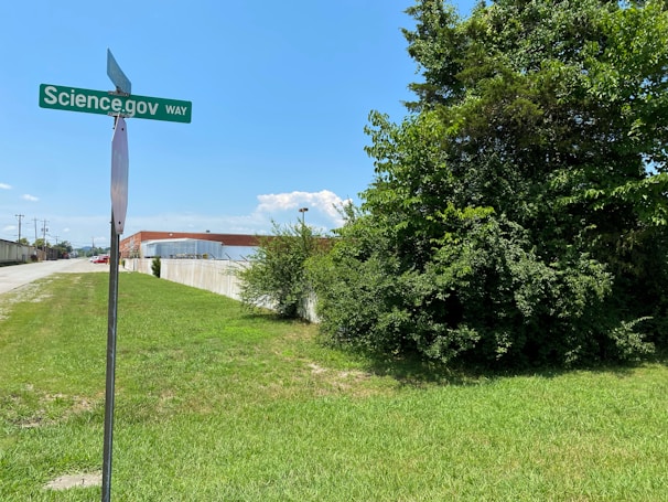 A street sign labeled 'Science.gov Way' stands in front of a grassy area beside a road. To the right, there is a large tree with dense green foliage. In the background, an industrial-looking building with a metal roof is visible beneath a clear blue sky.