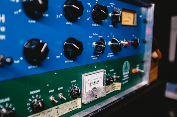 A close-up of an audio equipment panel with various knobs and switches. The top section is blue with dials labeled for audio control settings like ratio, threshold, and release. Below, a green section with additional controls and a vintage-style meter labeled 'TESLA'.
