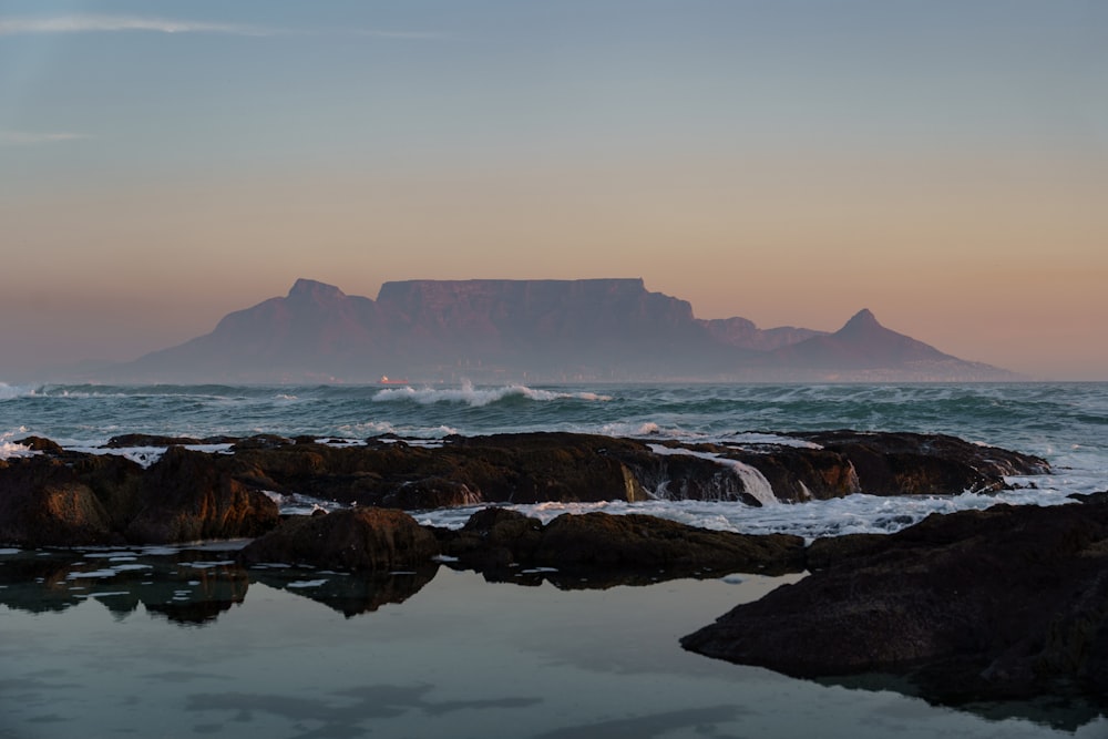 a rocky beach with a body of water and mountains in the background with Table Mountain in the background