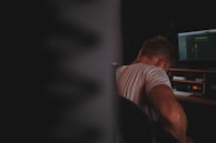 A person is sitting in a dimly lit room facing away from the camera, focusing on a computer screen displaying audio or video editing software. The person's back is dressed in a light-colored shirt and the workspace includes various electronic equipment.