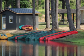 Several canoes and kayaks are lined up on the edge of a calm lake. The boats are primarily green and red in color. A small shed is nearby with more kayaks or canoes stacked beside it. Tall trees and a grassy area surround the buildings and the water. In the background, there is a picnic table under an open shelter.