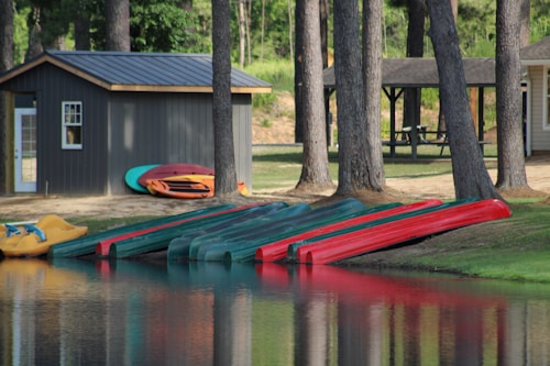 Several canoes and kayaks are lined up on the edge of a calm lake. The boats are primarily green and red in color. A small shed is nearby with more kayaks or canoes stacked beside it. Tall trees and a grassy area surround the buildings and the water. In the background, there is a picnic table under an open shelter.