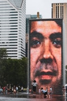 A large digital screen displays a close-up image of a face on a brick tower, situated in an urban environment surrounded by modern skyscrapers. People, including children, are playing and walking around the area, which appears to be a public space with water fountains.