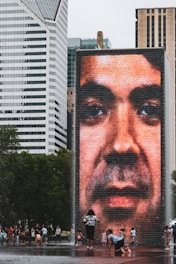 A large digital screen displays a close-up image of a face on a brick tower, situated in an urban environment surrounded by modern skyscrapers. People, including children, are playing and walking around the area, which appears to be a public space with water fountains.