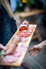 A wooden platter is being held by someone with slices of various cured meats including salami and sausages. The focus is on the arranged assortment with a blurred hand reaching out towards it. The background is slightly out of focus, featuring a table and some greenery.