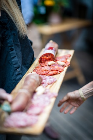 A wooden platter is being held by someone with slices of various cured meats including salami and sausages. The focus is on the arranged assortment with a blurred hand reaching out towards it. The background is slightly out of focus, featuring a table and some greenery.
