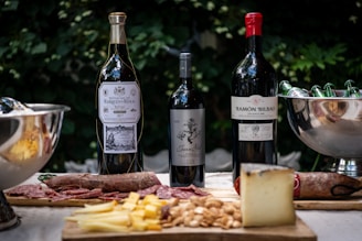 A table set with three bottles of wine as the central focus, accompanied by charcuterie including sliced meats, cheese, and nuts. A silver champagne bucket filled with bottled drinks is on the side. The backdrop features lush green foliage, creating an inviting and leisurely outdoor dining atmosphere.