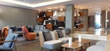 The image depicts a modern, stylishly decorated living room or lounge area with contemporary furniture. There are various seats, including sofas, armchairs, and ottomans, arranged in a cozy and inviting manner. Shelving units in the background display decorative items. The space is well-lit with both natural and artificial light, creating a warm and welcoming atmosphere.