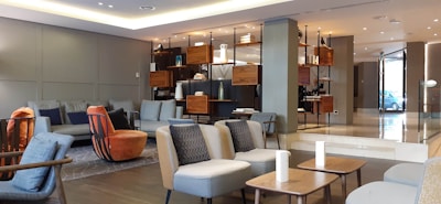 The image depicts a modern, stylishly decorated living room or lounge area with contemporary furniture. There are various seats, including sofas, armchairs, and ottomans, arranged in a cozy and inviting manner. Shelving units in the background display decorative items. The space is well-lit with both natural and artificial light, creating a warm and welcoming atmosphere.