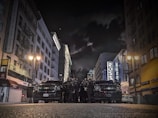 A city street at night with high-rise buildings lining both sides, illuminated by streetlights. A group of law enforcement officers is gathered in front of two black vehicles in the middle of the street.