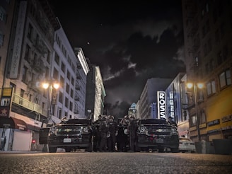A city street at night with high-rise buildings lining both sides, illuminated by streetlights. A group of law enforcement officers is gathered in front of two black vehicles in the middle of the street.