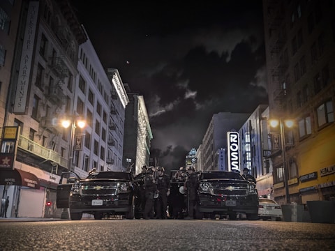 A city street at night with high-rise buildings lining both sides, illuminated by streetlights. A group of law enforcement officers is gathered in front of two black vehicles in the middle of the street.