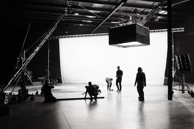 A well-lit film or photography studio setting with several people working. The backdrop is an expansive white screen with lighting equipment overhead and on the sides. Individuals are scattered throughout, some appearing to adjust equipment or collaborate on tasks.