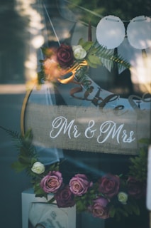 A decorative arrangement featuring a sign with 'Mr & Mrs' written on burlap fabric, surrounded by an array of artificial flowers in soft pinks, purples, and whites. The flowers, along with green leaves, form a garland, contributing to the romantic and elegant setting. Light reflections are visible on the glass surface, adding a dreamy quality to the scene.
