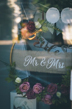 A decorative arrangement featuring a sign with 'Mr & Mrs' written on burlap fabric, surrounded by an array of artificial flowers in soft pinks, purples, and whites. The flowers, along with green leaves, form a garland, contributing to the romantic and elegant setting. Light reflections are visible on the glass surface, adding a dreamy quality to the scene.