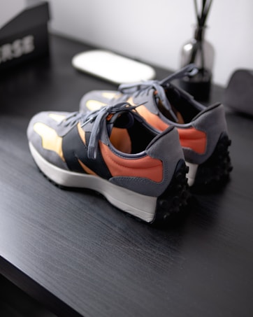 A pair of sneakers with grey, black, peach, and beige colors are placed on a dark wooden surface. The sneakers have a modern design with a white sole and textured black grip at the heel. Near the shoes, there is a white object that appears to be a mousepad or a small tray, as well as a reflective bottle with sticks protruding from the top, likely a reed diffuser. Part of a black box with white text is visible in the background.