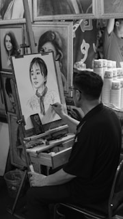 A man is seated at an easel, drawing a portrait of a young woman in a detailed black-and-white sketch. The workspace is surrounded by other framed portraits, suggesting an art studio or gallery. Cups are stacked on the right, and a smartphone is placed on the easel.