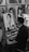 A man is seated at an easel, drawing a portrait of a young woman in a detailed black-and-white sketch. The workspace is surrounded by other framed portraits, suggesting an art studio or gallery. Cups are stacked on the right, and a smartphone is placed on the easel.