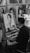 A man is seated at an easel, drawing a portrait of a young woman in a detailed black-and-white sketch. The workspace is surrounded by other framed portraits, suggesting an art studio or gallery. Cups are stacked on the right, and a smartphone is placed on the easel.