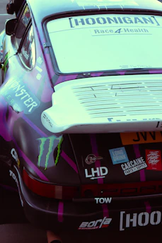 A close-up view of the rear section of a race car featuring various decals and stickers, including brand logos and racing team graphics. The vehicle's aesthetics are enhanced with a combination of dark purple and black colors with a prominent rear spoiler.