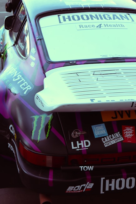 A close-up view of the rear section of a race car featuring various decals and stickers, including brand logos and racing team graphics. The vehicle's aesthetics are enhanced with a combination of dark purple and black colors with a prominent rear spoiler.