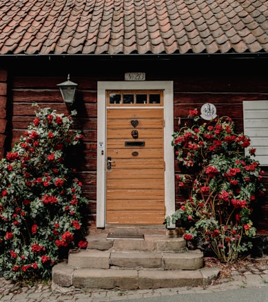 A rustic wooden front door is surrounded by vibrant red flowers and green foliage. The entryway features a stone step and cobblestone path, with a brick wall exterior and a classic lantern-style light fixture beside the door.