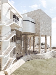 A modern architectural structure featuring a blend of stone and glass materials. The building has multiple levels accessed by external staircases with metal railings. Tall columns support the upper structure, and there are areas with outdoor seating. The architecture includes clean, minimalist lines and surfaces that create an aesthetic contrast between natural stone and sleek glass.