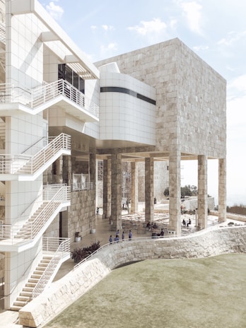 A modern architectural structure featuring a blend of stone and glass materials. The building has multiple levels accessed by external staircases with metal railings. Tall columns support the upper structure, and there are areas with outdoor seating. The architecture includes clean, minimalist lines and surfaces that create an aesthetic contrast between natural stone and sleek glass.