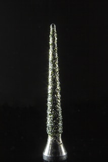 A metallic, conical object stands vertically against a dark background. Its surface is textured with a rough, clustered pattern that reflects light, creating a sparkling effect.