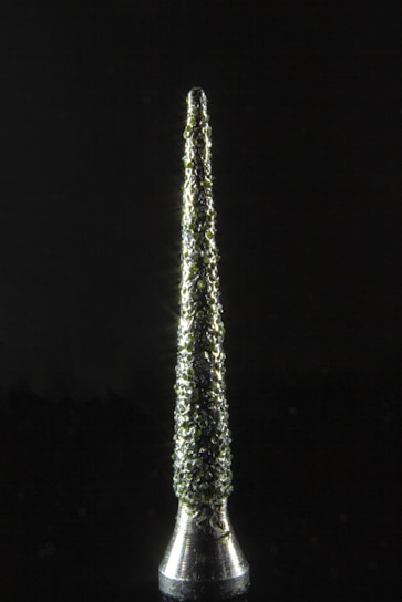 A metallic, conical object stands vertically against a dark background. Its surface is textured with a rough, clustered pattern that reflects light, creating a sparkling effect.