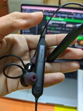 A hand is holding a pair of in-ear wireless earphones with a black cable and pink tips. The brand 'OnePlus' is visible on the neckband. In the background, there is a blurred view of a laptop screen displaying colorful graphics and text.